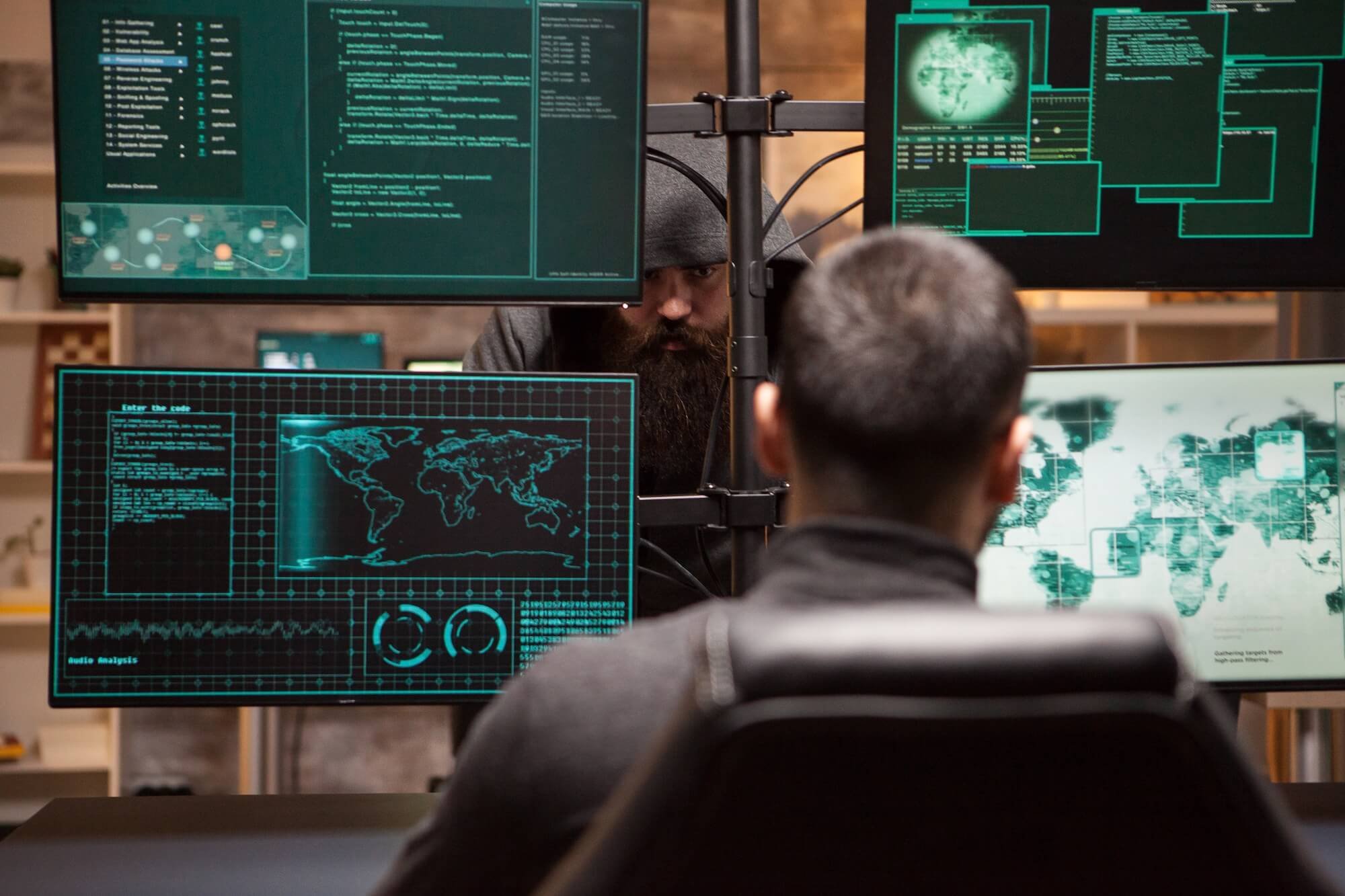 Two people working on multiple computer screens displaying global maps, data, and code in a high-tech environment.
