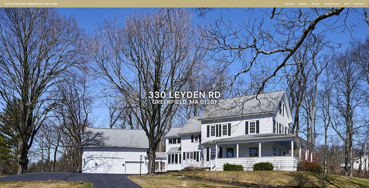 Large two-story white house with a wrap-around porch, set on a grassy hill with leafless trees in the background on a clear day.