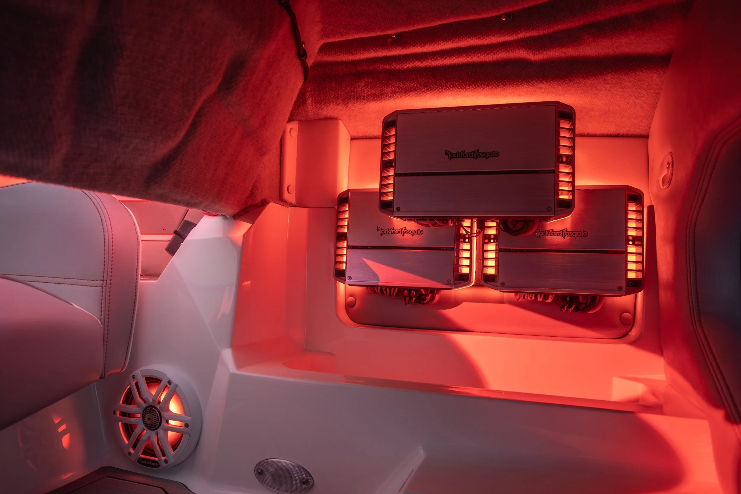 The image shows three Rockford Fosgate car audio amplifiers installed on a beige or light-colored panel in the interior of a vehicle, illuminated with red lighting.