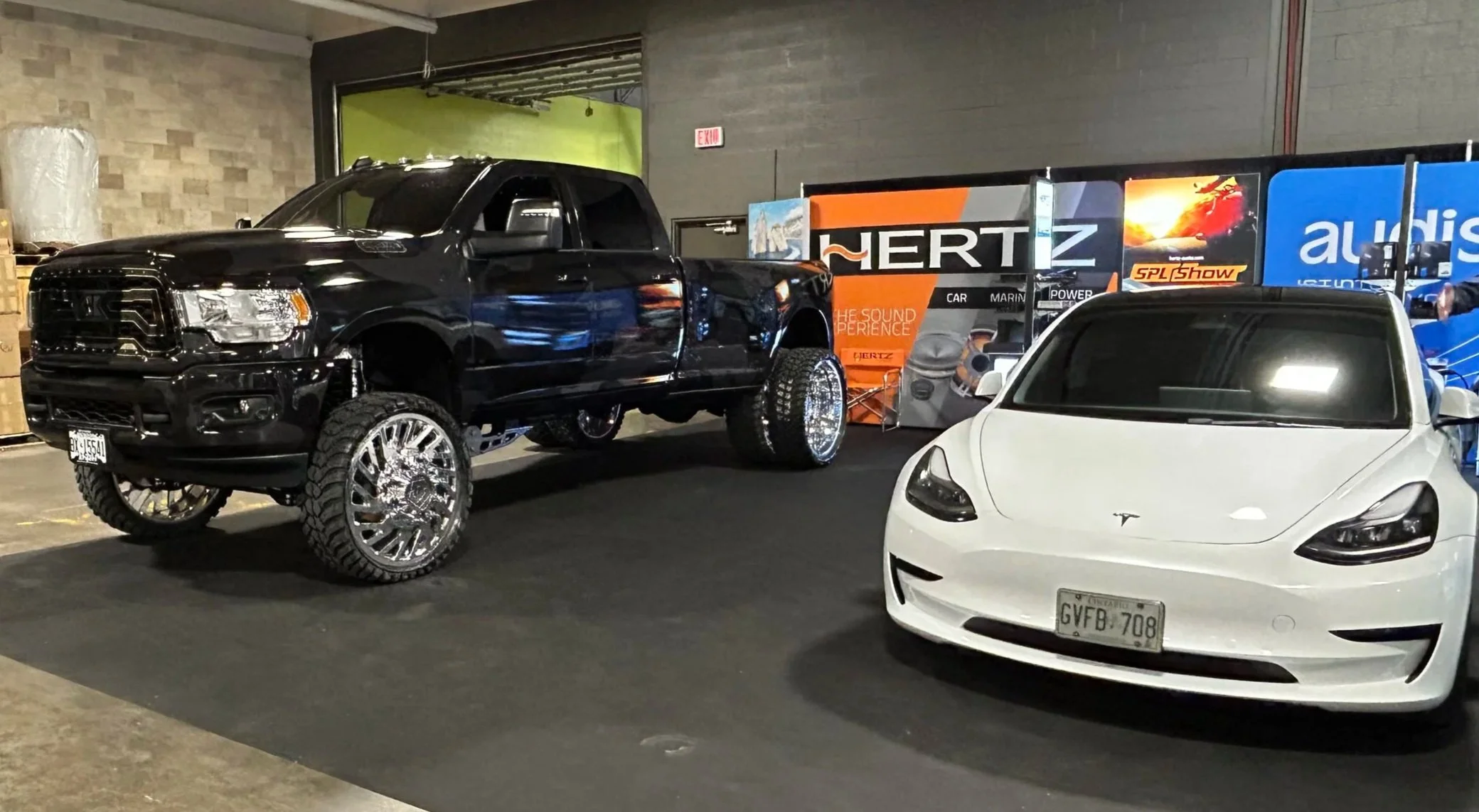 A black lifted pickup truck with chrome wheels and large off-road tires parked indoor next to a white Tesla Model 3 with tinted windows. In the background, there are banners and signs including 'HERTZ' and 'Audison'.