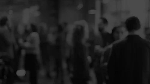 Blurred image of a group of people standing and socializing in a dimly lit room.