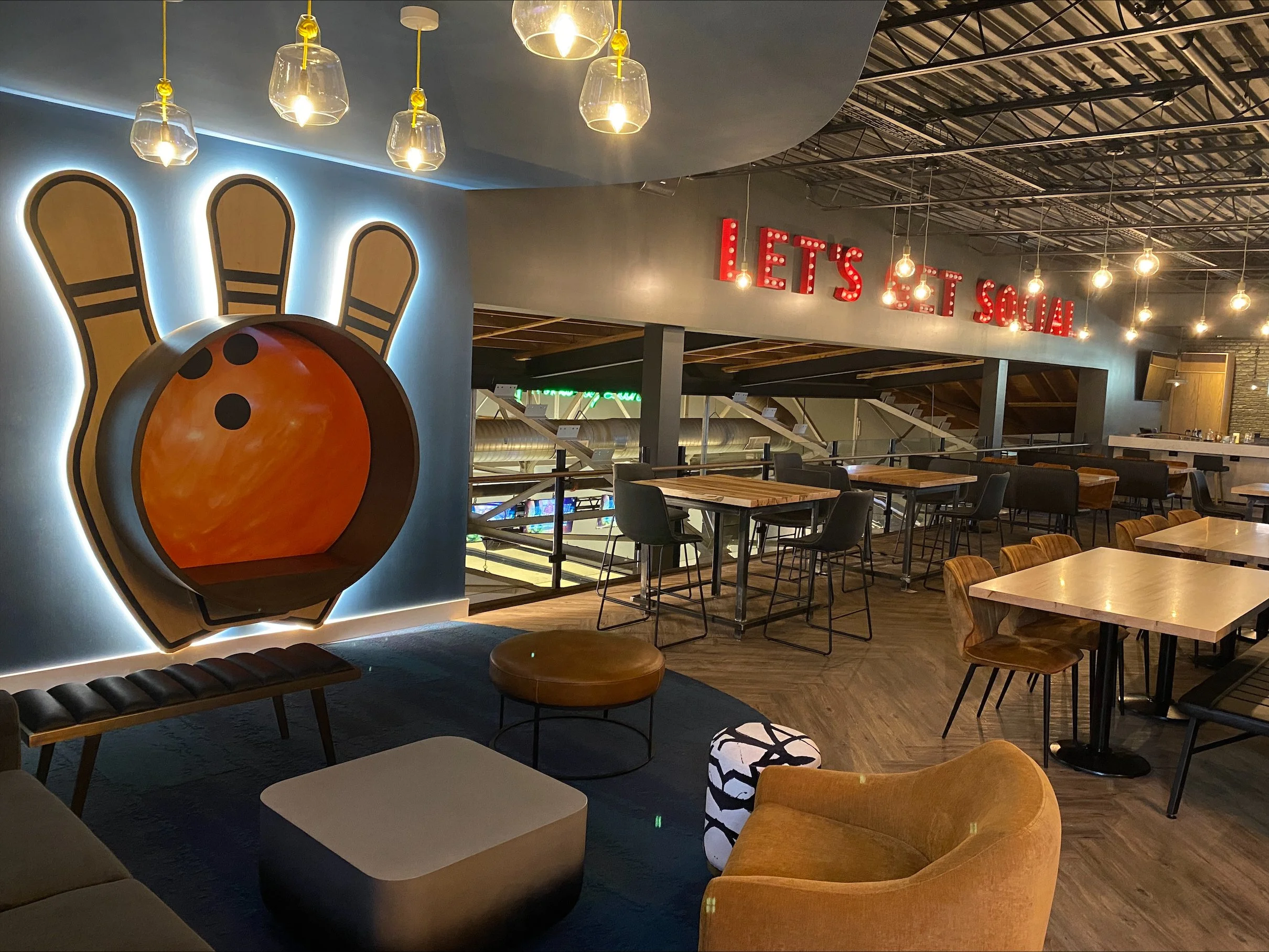 Interior of a social lounge at a bowling alley featuring a large bowling ball and pins wall art, with seating areas, tables, and a neon sign that reads 'LET'S SOCIAL'.