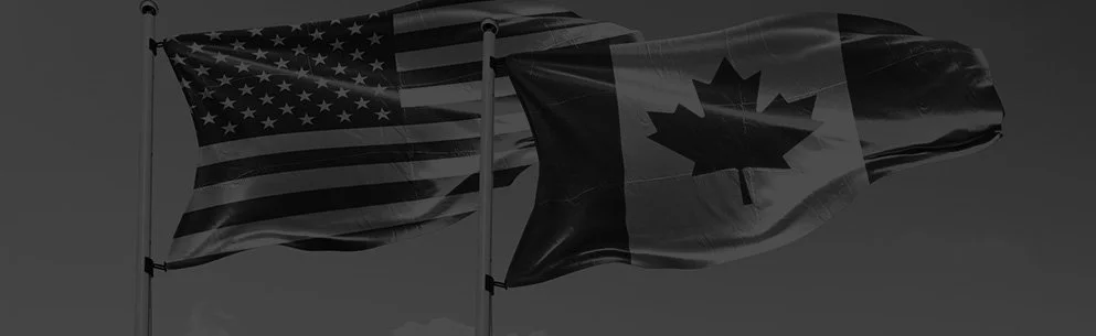 Two flags waving in the wind, one American flag and one Canadian flag with a maple leaf symbol, against a cloudy sky.