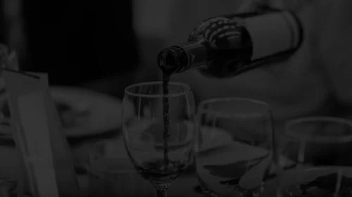 A bottle pouring wine into a wine glass on a table, with other glasses and bottles visible in the background.