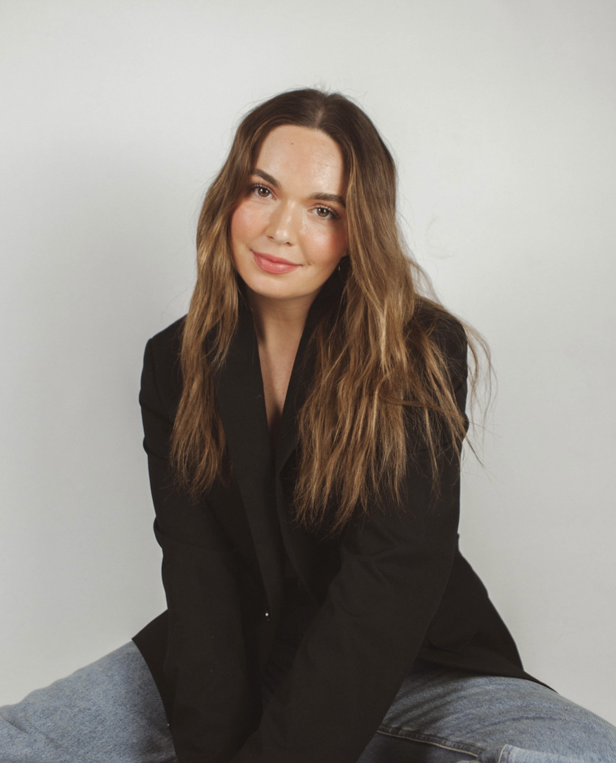 A woman with long, wavy brown hair wearing a black blazer and gray jeans, sitting against a plain white background.