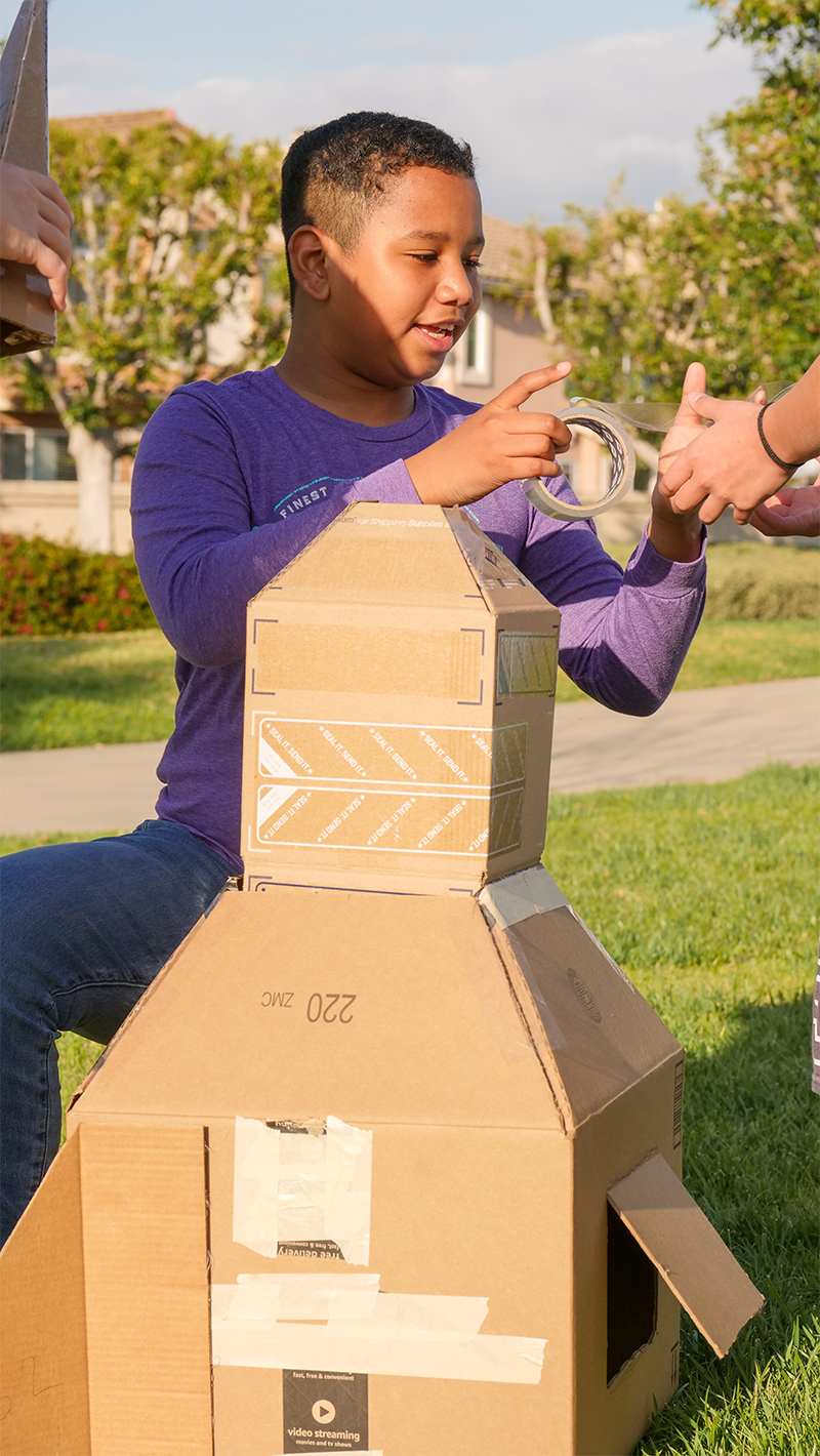 A boy wearing a purple long sleeve shirt stacking cardboard boxes outdoors while holding packing tape, with trees and houses in the background.