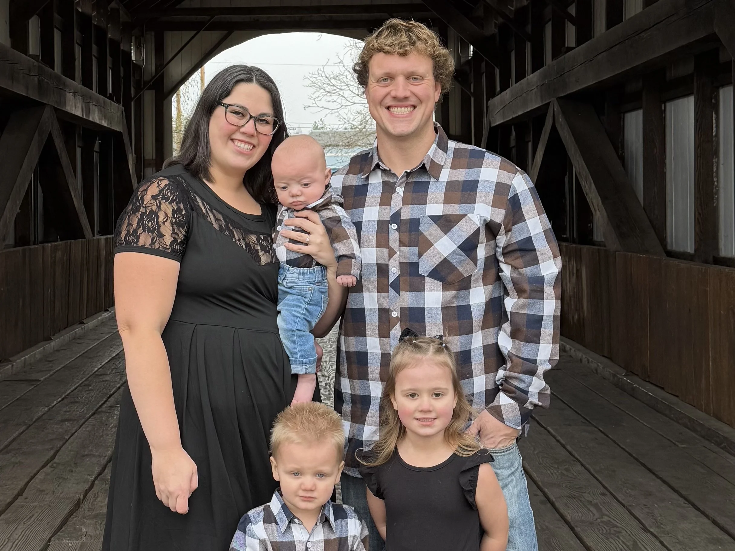 A happy family of four outdoors in front of green bushes, with a man, woman, and two young children, all smiling.