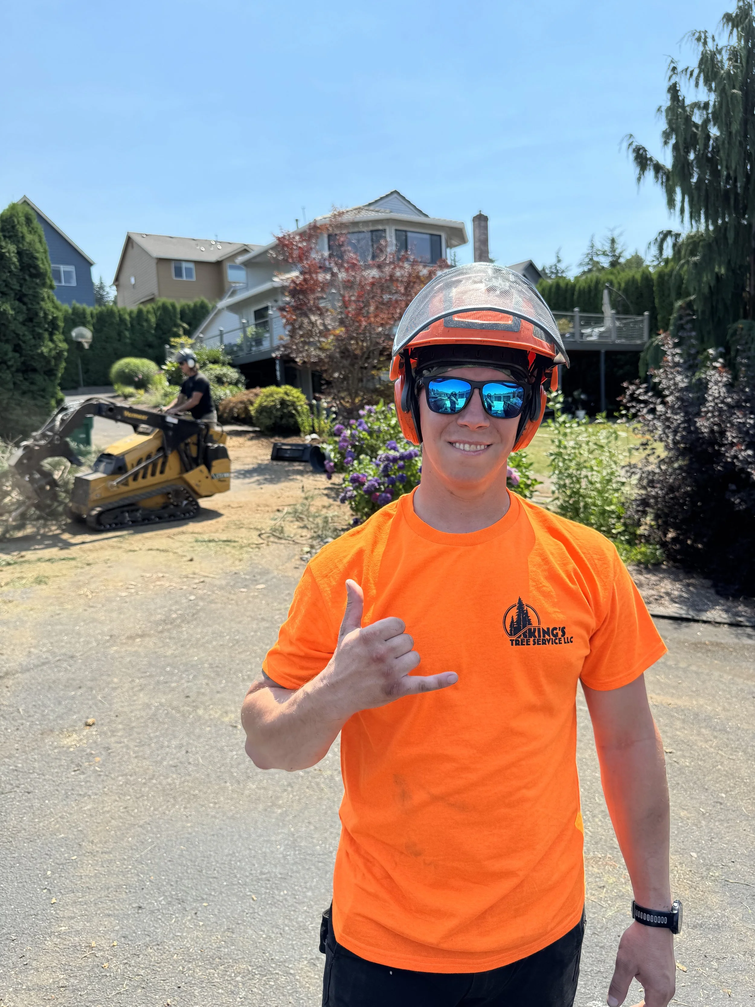 A smiling man dressed in an orange t-shirt, sunglasses, and a helmet giving a thumbs-up gesture. In the background, a person is operating a small construction vehicle, and there are houses and trees in a suburban yard.