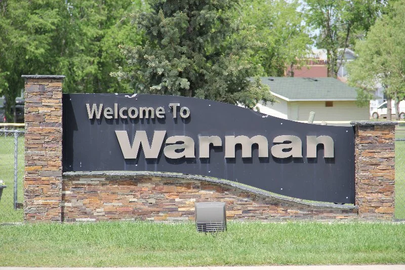A large black sign with gray and white lettering that reads "Welcome to Warman". The sign is supported by stone pillars on each side, situated on a grassy area with trees and houses in the background.