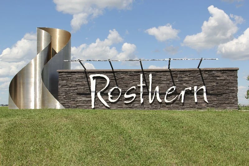 Sign with the word 'Rosthern' on a stone wall, with a metallic spiral sculpture to the left and a cloudy blue sky overhead.