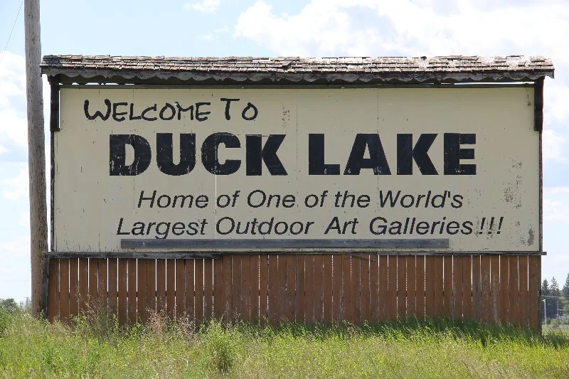 A large outdoor sign welcoming visitors to Duck Lake, advertising it as home to one of the world's largest outdoor art galleries, with a cloudy sky and grass in the foreground.