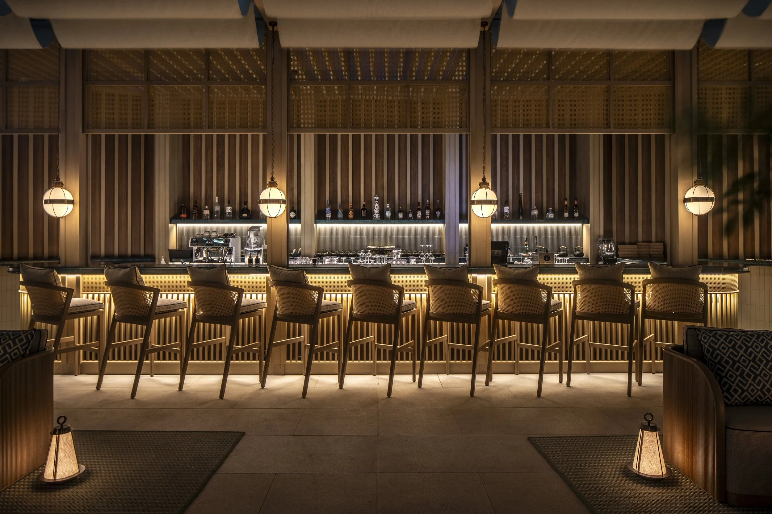The outdoor bar at Tattu Dubai Sky Pool at nighttime. Rattan bar stool and softly glowing lanterns are in the foreground.