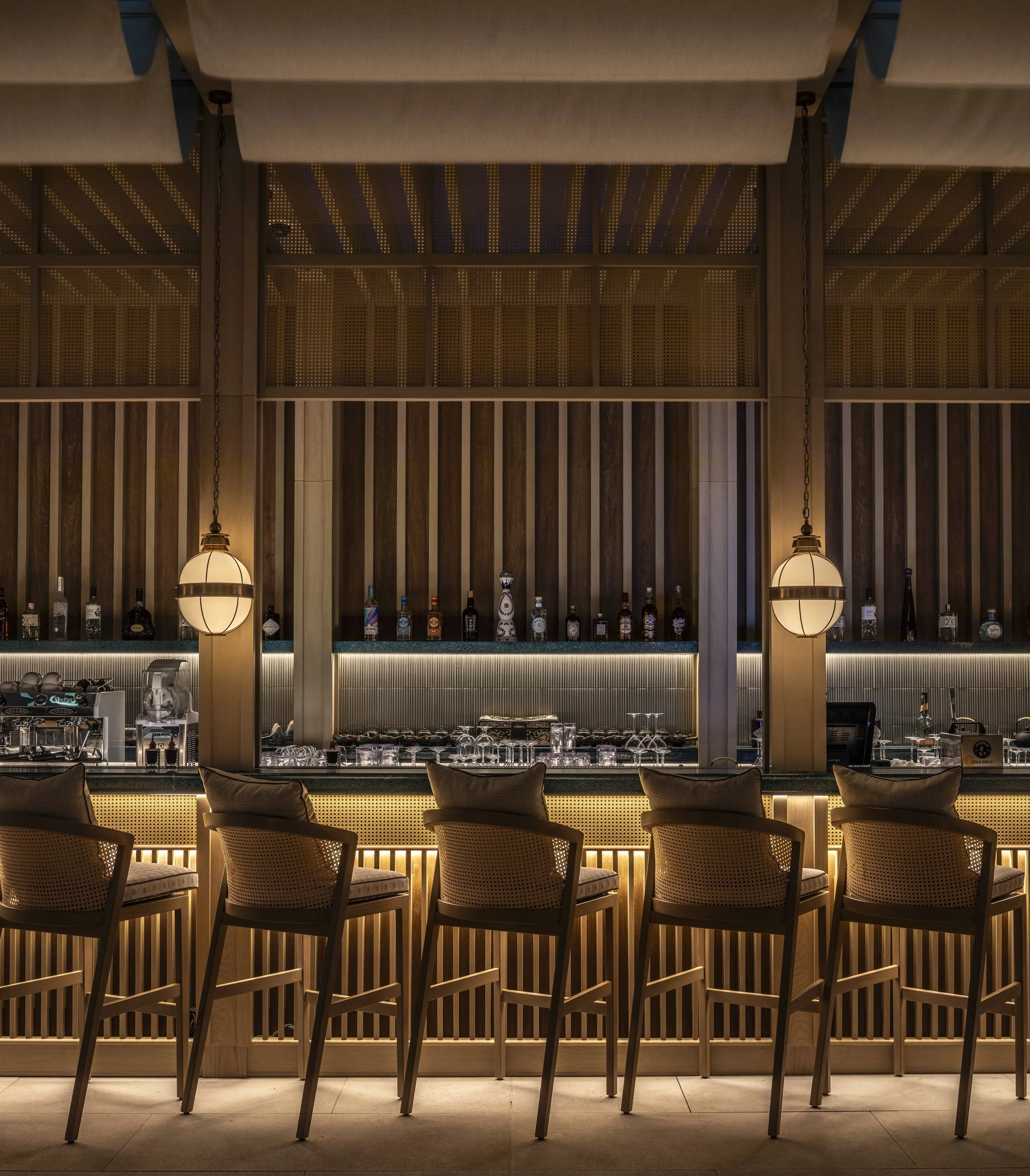 The outdoor bar on the pool deck, with light timber slatted detail and rough tiles in the background. Rattan backed bar stools match the rattan headers.