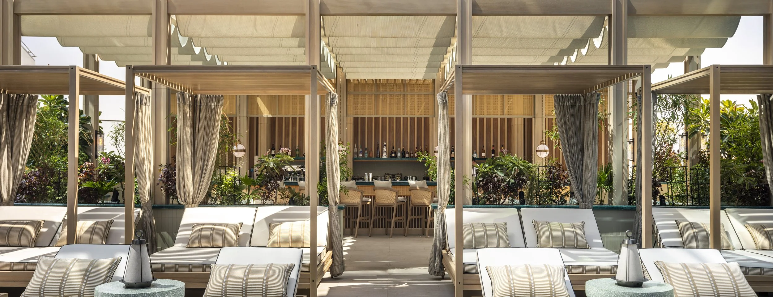 A view of the pool deck wide angle with loungers in the foreground, cabanas in the middle and behind a screen of planting there is a cocktail bar.