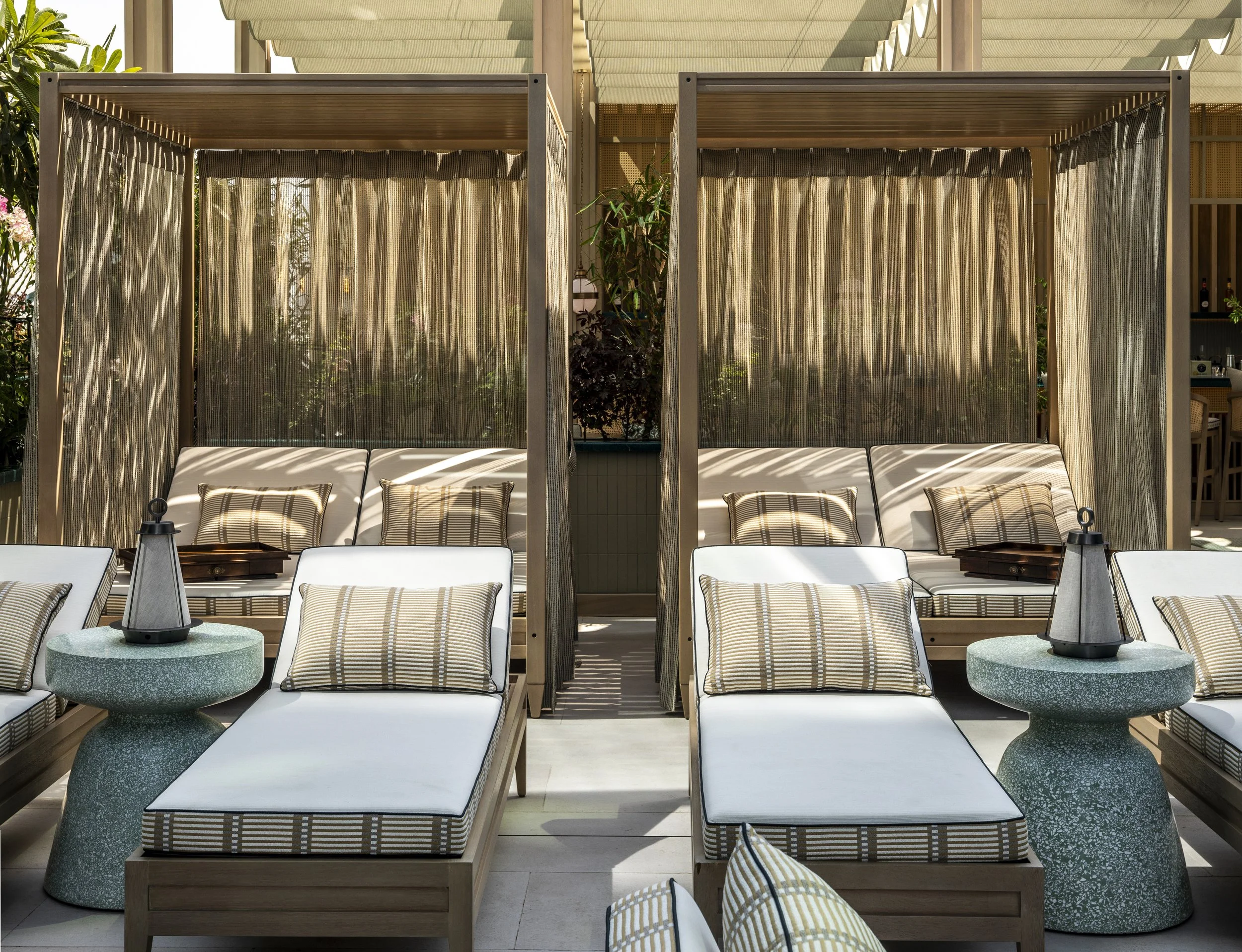 Luxury outdoor lounge area with cabana-style seating, white cushioned daybeds, striped pillows, round side tables with lanterns, and natural sunlight.