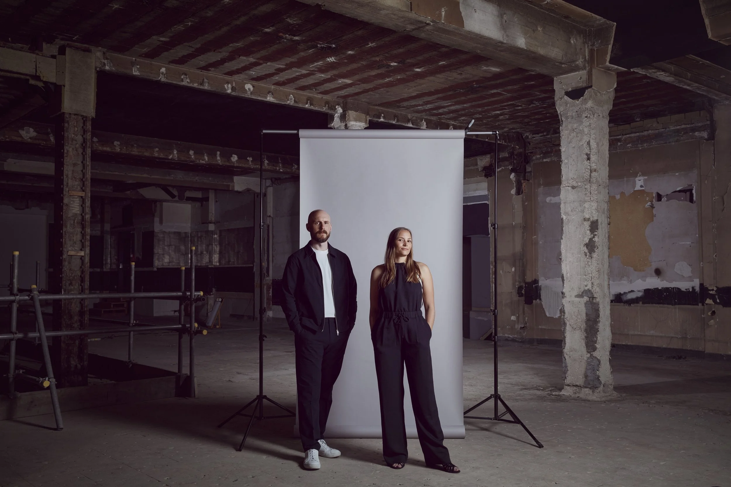 Chris and Christina, co-founders and principals of Studio WYZE, standing in front of a white backdrop within the concrete shell of their FENIX Mayfair site. Unfinished space with exposed concrete and brick walls.