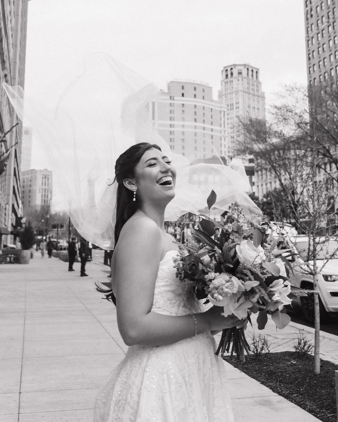 Candid black and white photo of a bride laughing as the wind blows her veil in downtown Detroit.