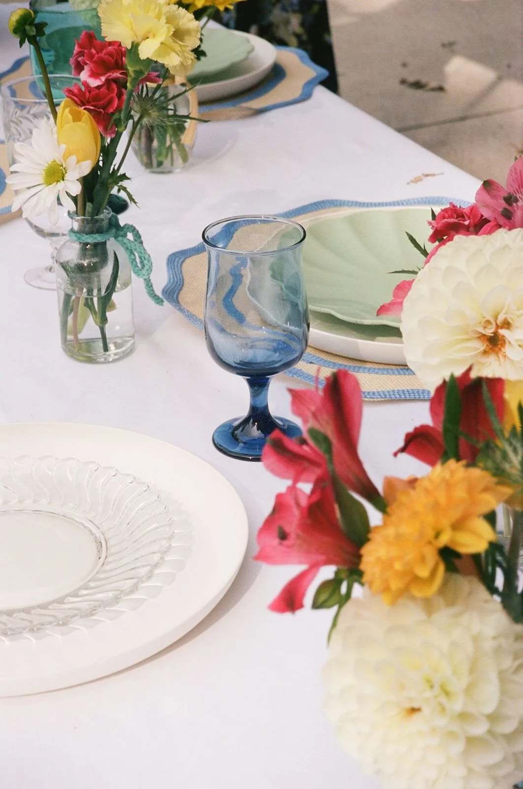 A 35mm film photo of a backyard wedding tablescape. There are thrifted and curated plates and glasses on the white linen tablecloth.