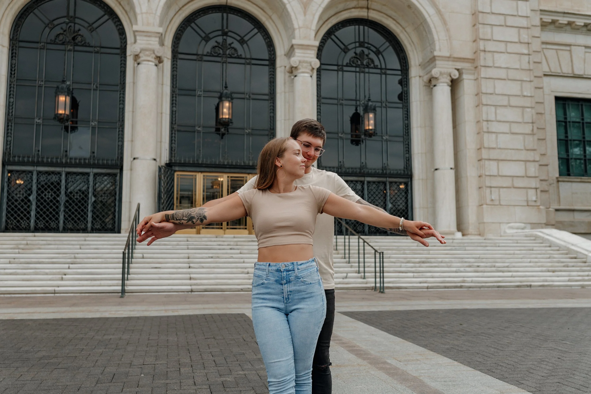 A young couple standing in front of the steps at the Detroit Institute of Art. He stands behind her and guides their arms into an airplane motion.