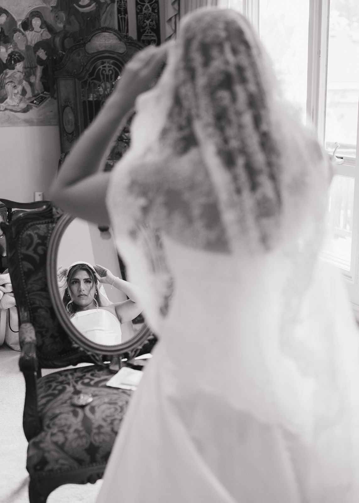 A black and white photo of a bride getting ready, taken through a mirror. She adjusts her veil in the mirror.