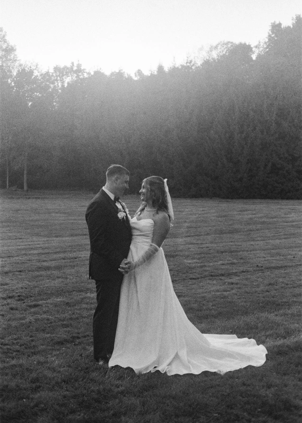 A classic full-body portrait of a bride and groom on black and white film. The stand side by side with their hands linked at their hips.