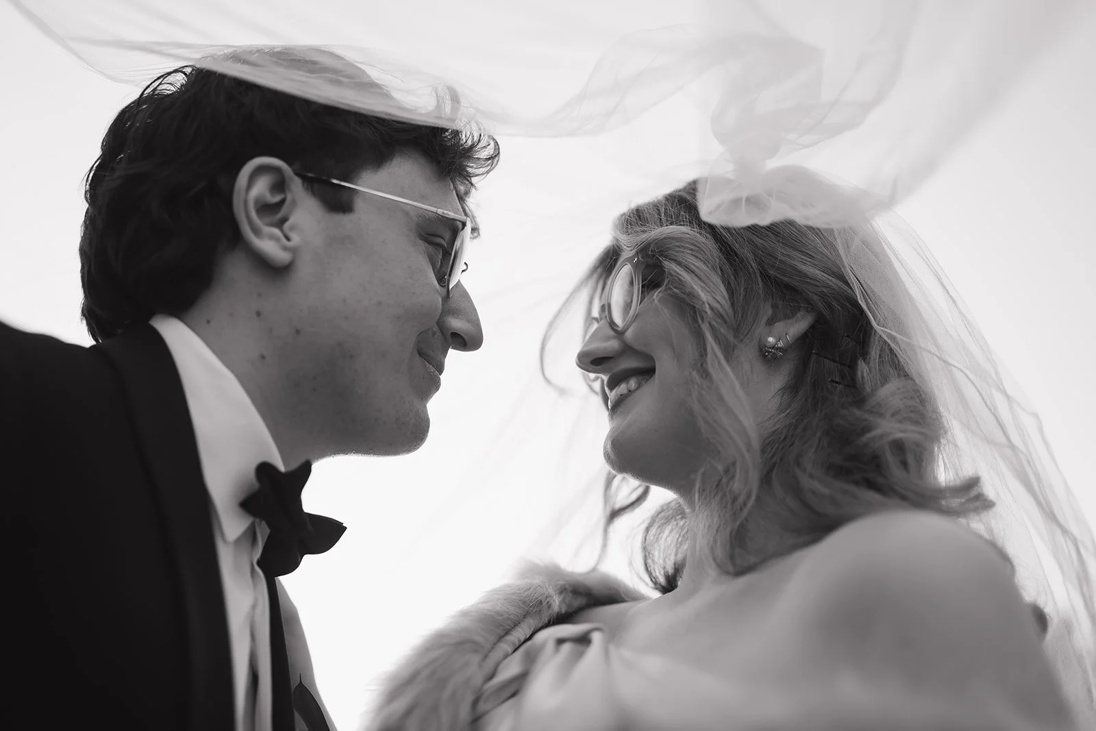Black and white portrait of a bride and groom smiling at each other under the brides veil.