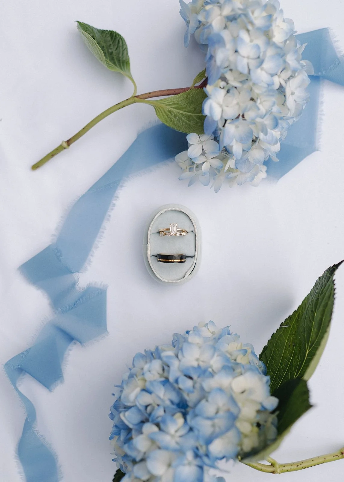 A detail shot of a bride and groom's wedding bands in a blue velvet ring box. Blue hydrangeas encircle the ring box, and a blue ribbon lies underneath.
