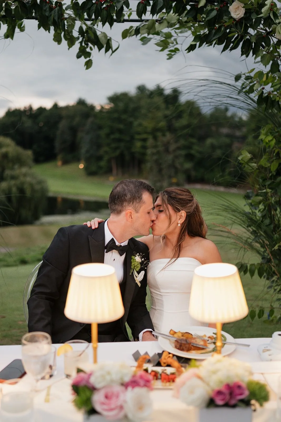 A newly-wedded couple kissing under the floral arch at their sweetheart table at dusk.
