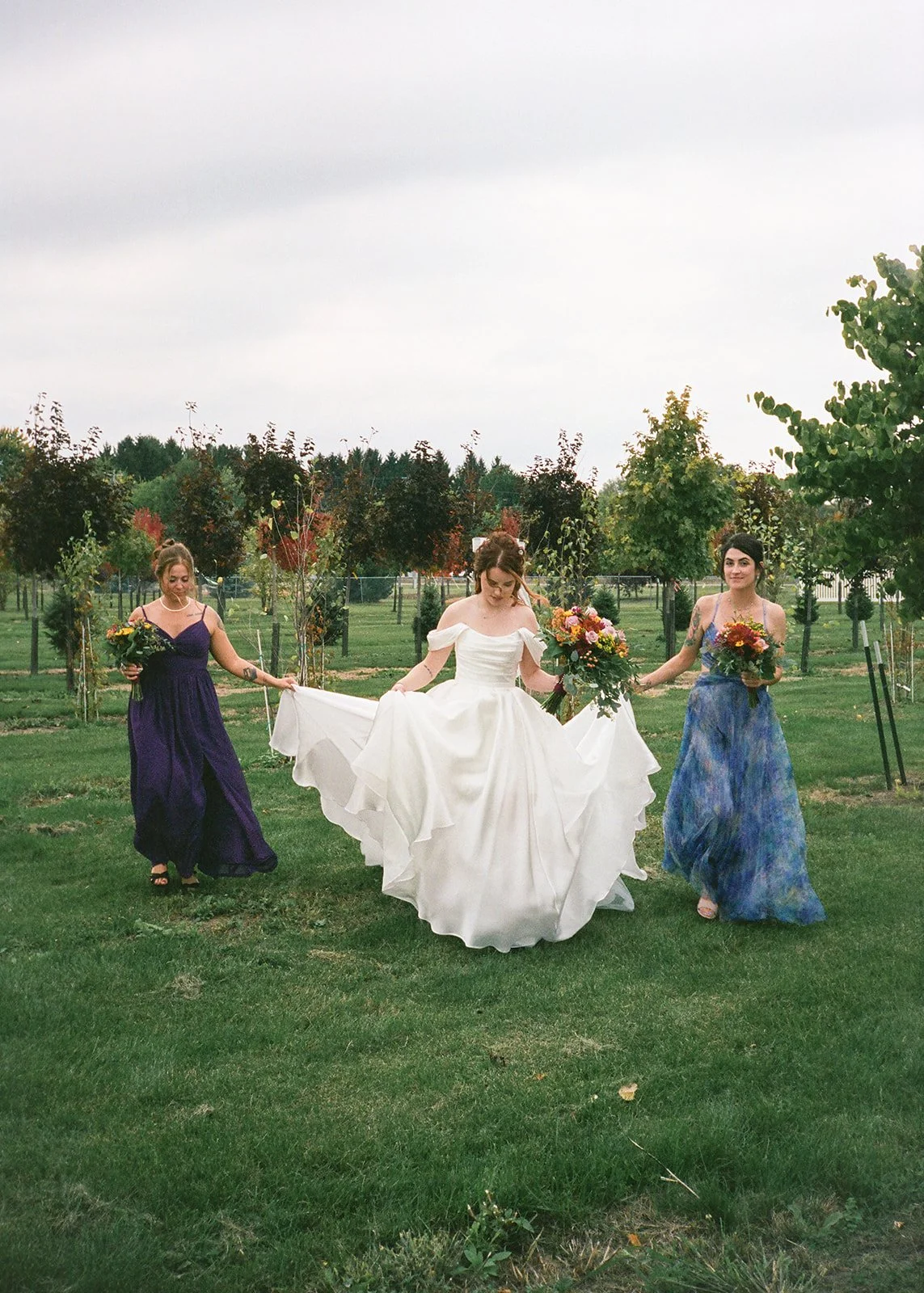 Film photo of bridesmaids holding the brides train as she walks in the grass.