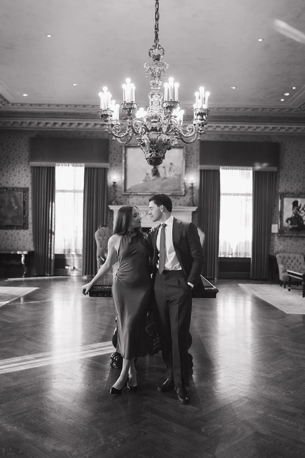 Black and white photo of a couple standing beneath a chandelier at The Detroit Athletic Club for their engagement photos.