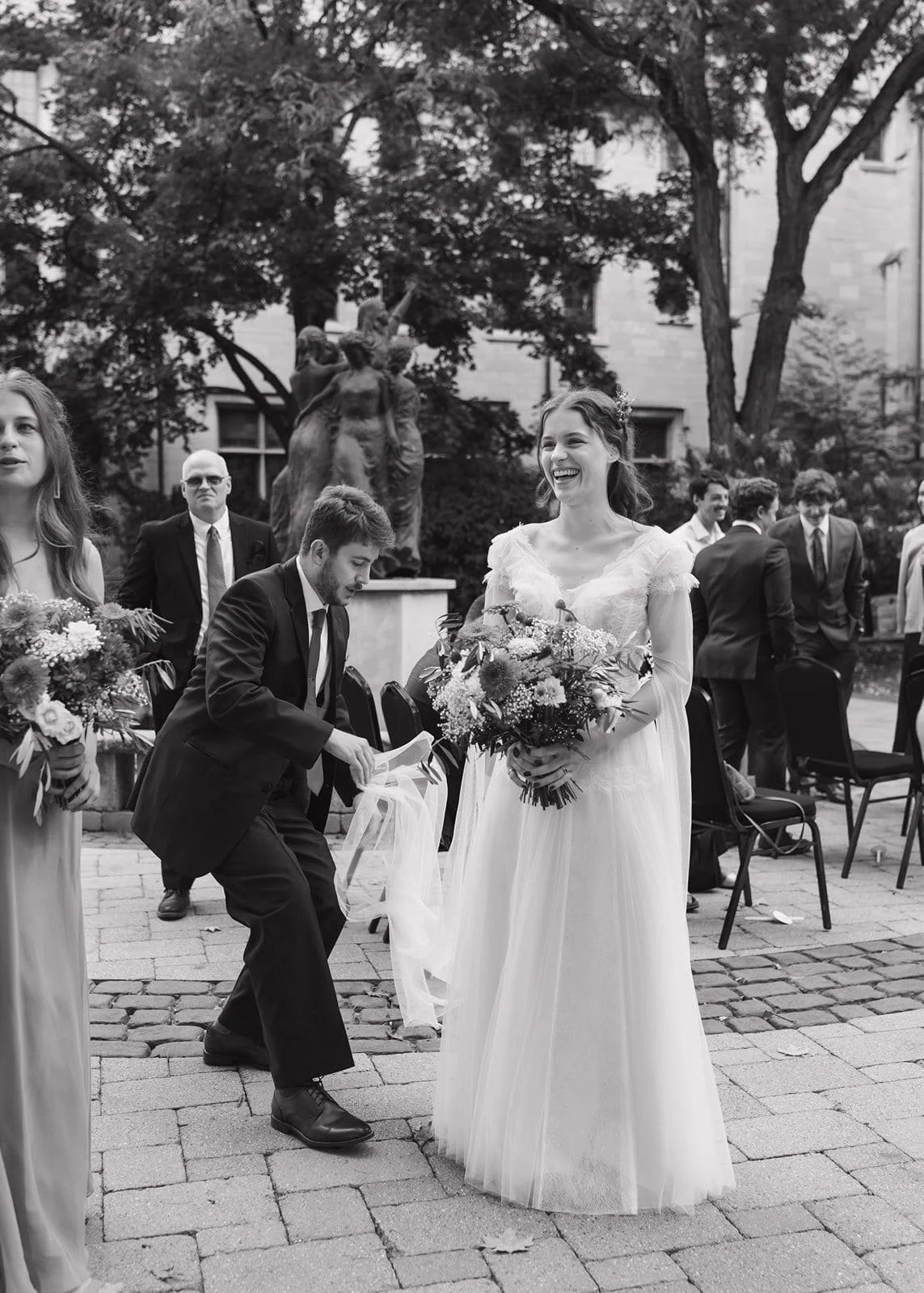 A black and white photo of a groom helping his new wife carry her dress after their ceremony at Marygrove Conservancy in Detroit, Michigan.