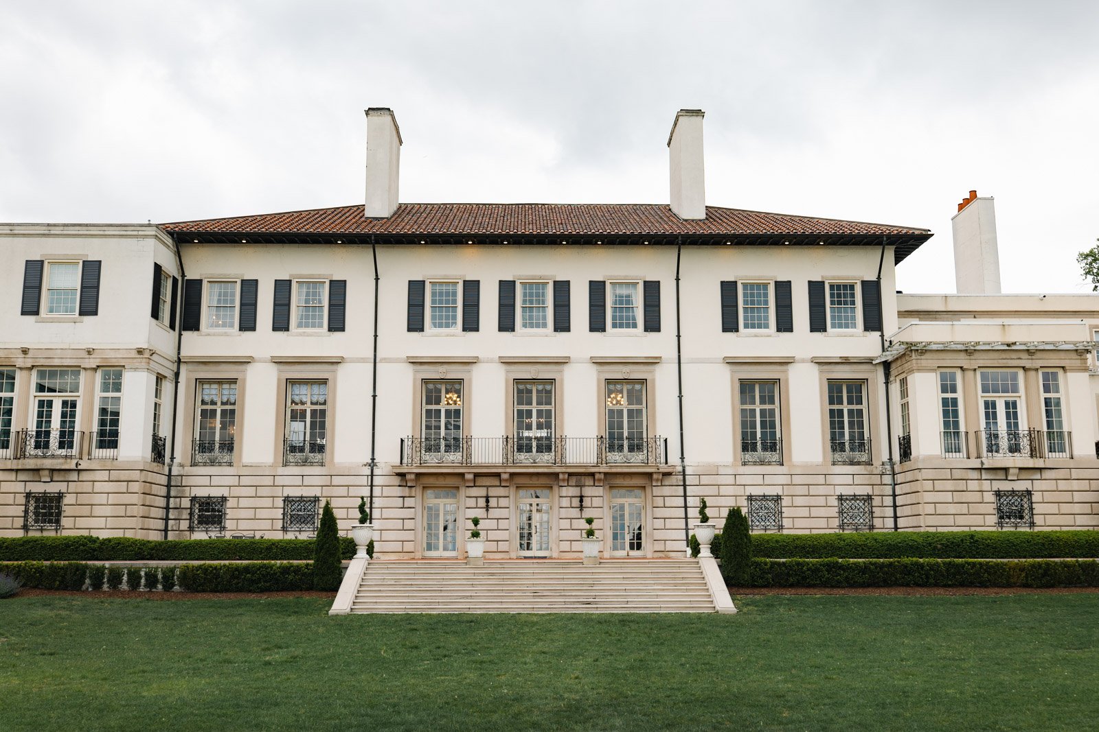 An exterior photo of the back grounds of the War Memorial wedding venue. It is a light tan colored building with red tiles on the roof, green hedges and lush green grass for outdoor wedding ceremonies.