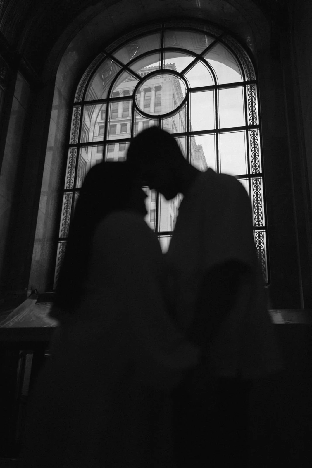 A black and white photo of two silhouettes standing forehead to forehead. There is a tall, arched window in the background outlining their figures.