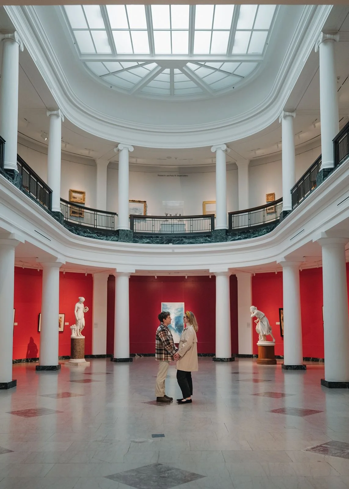 Art museum engagement photos of a couple standing together inside University of Michigan Museum of Art with red gallery walls and classical architecture