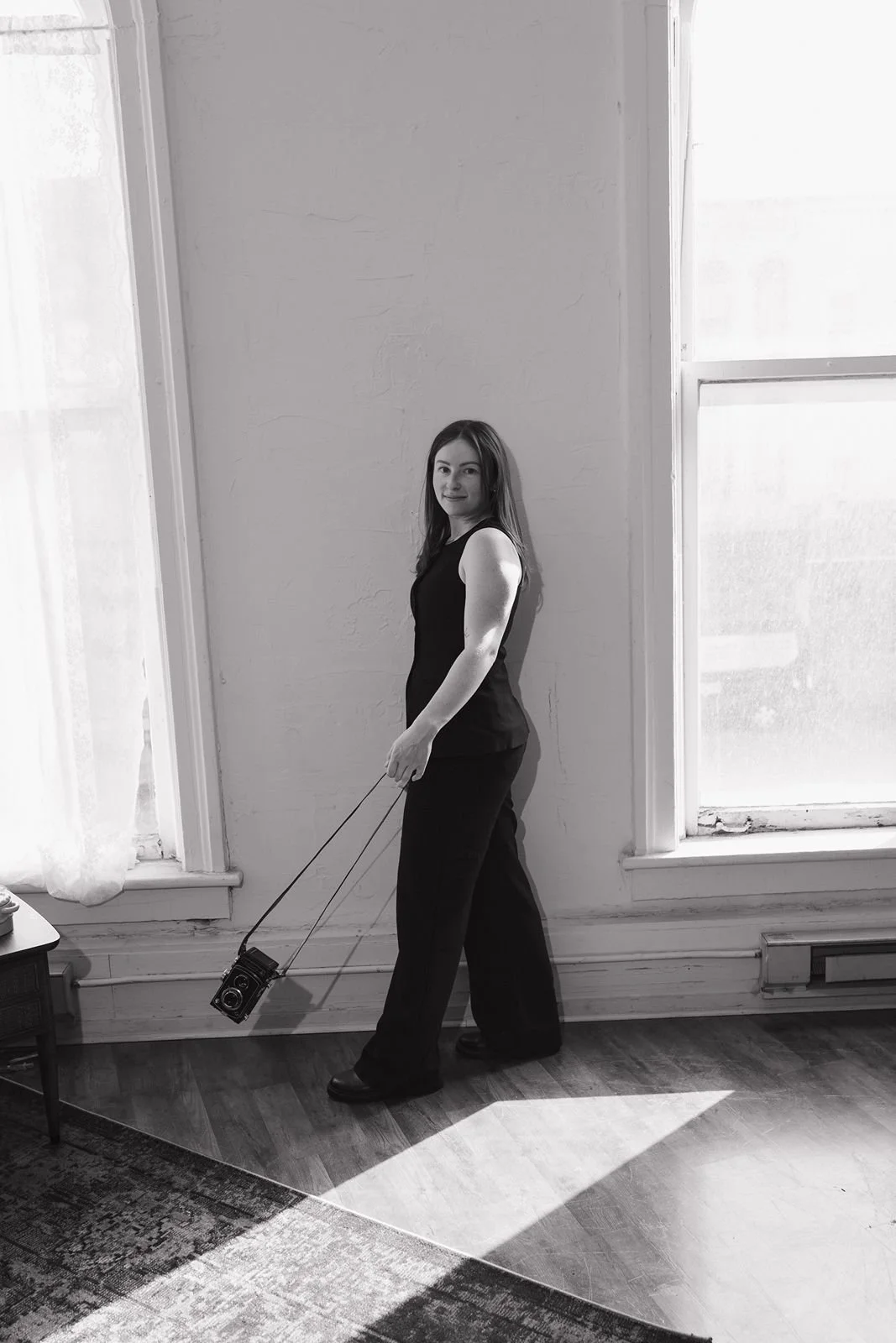 A headshot of Alaina, a Detroit Wedding Photographer , wearing a black pant suit against a white backdrop, swinging a 120 film camera..