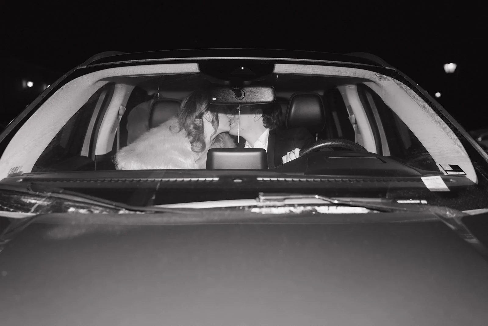Black and white photo of a couple kissing inside their getaway wedding car.
