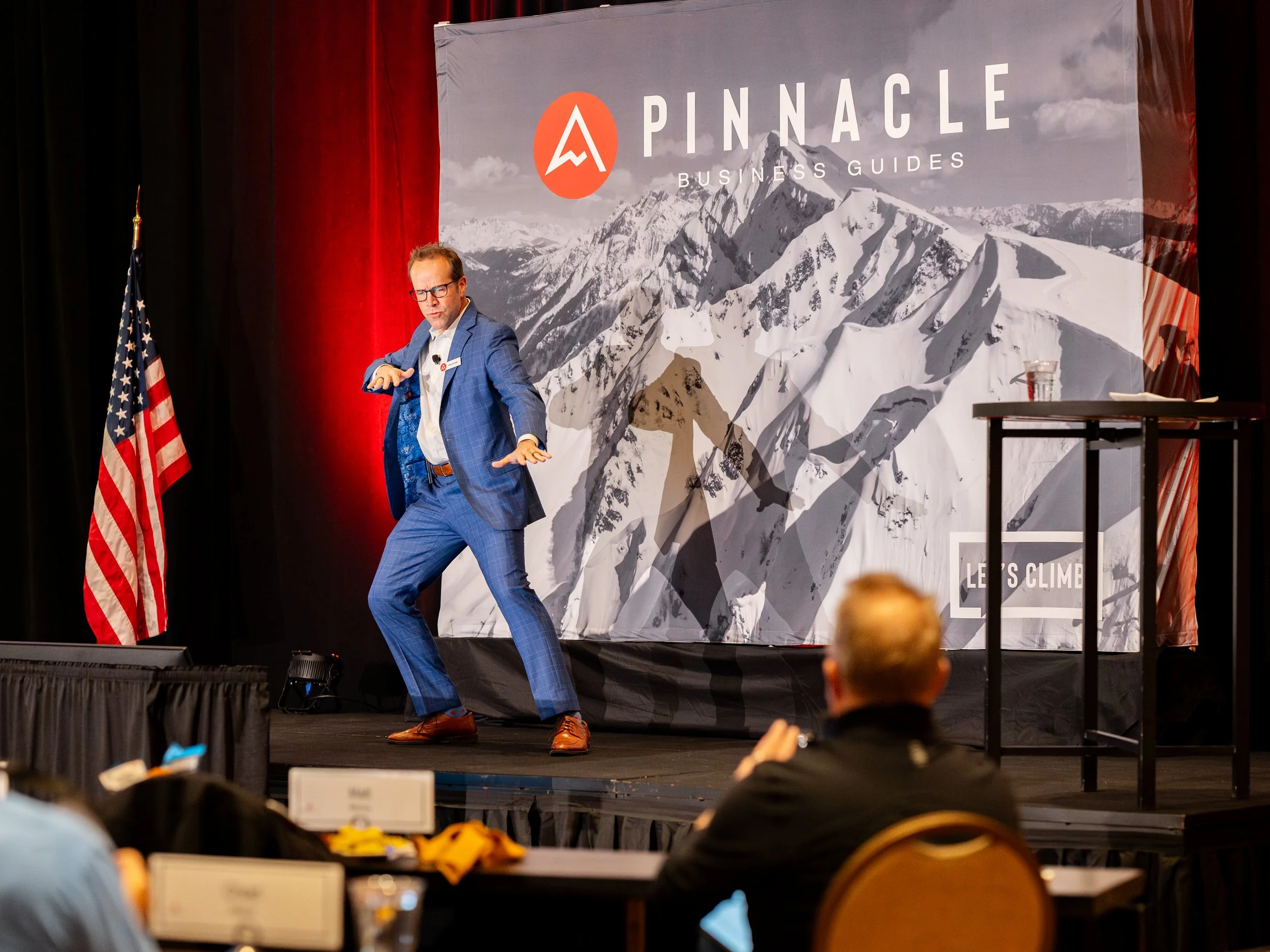 A man in a blue checkered suit dancing or performing on stage with a mountain-themed backdrop that says 'Pinnacle Business Guides.' An American flag stands to the left, and an audience member is visible in the foreground watching the performance.