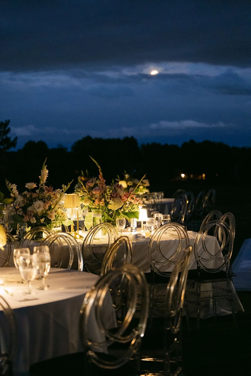 A photo of an outdoor wedding reception space at dusk, with golden glowing lamps and floral arrangements on the table.