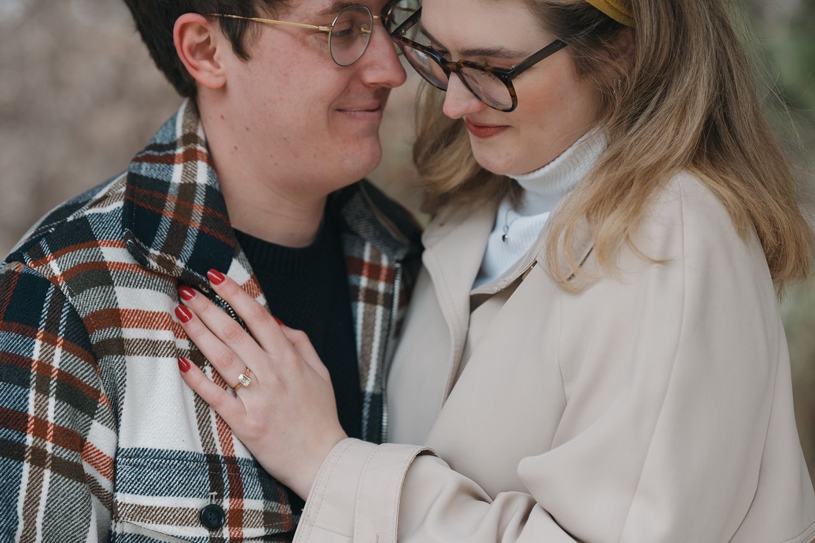 Close-up of engaged couple in Ann Arbor sharing an intimate moment, woman showing engagement ring while wearing neutral trench coat and headband