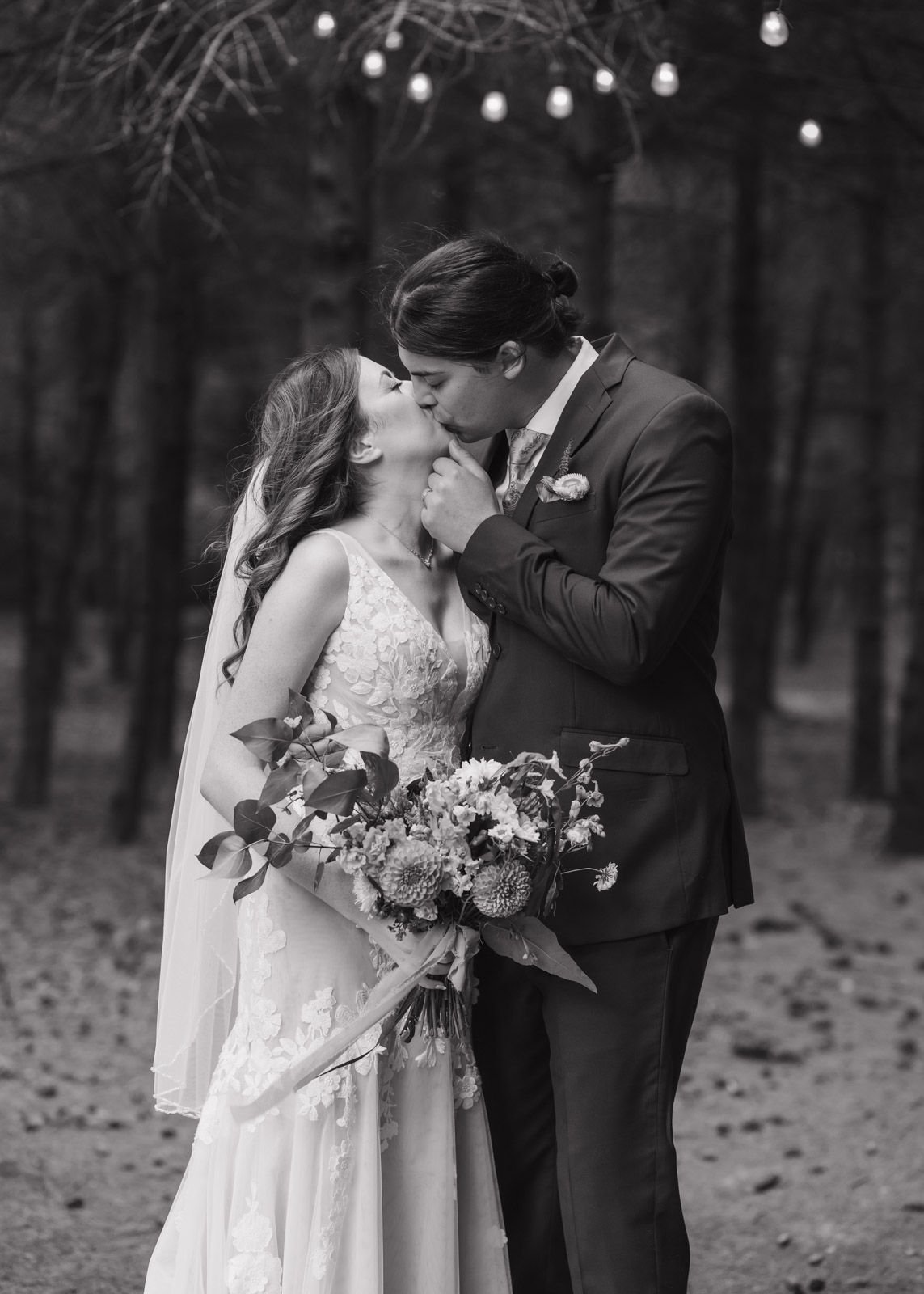 A black and white photo of a newly-married couple sharing a kiss. His hand is on her chin as she dips her head up to meet him. There are pine trees and string lights in the background.
