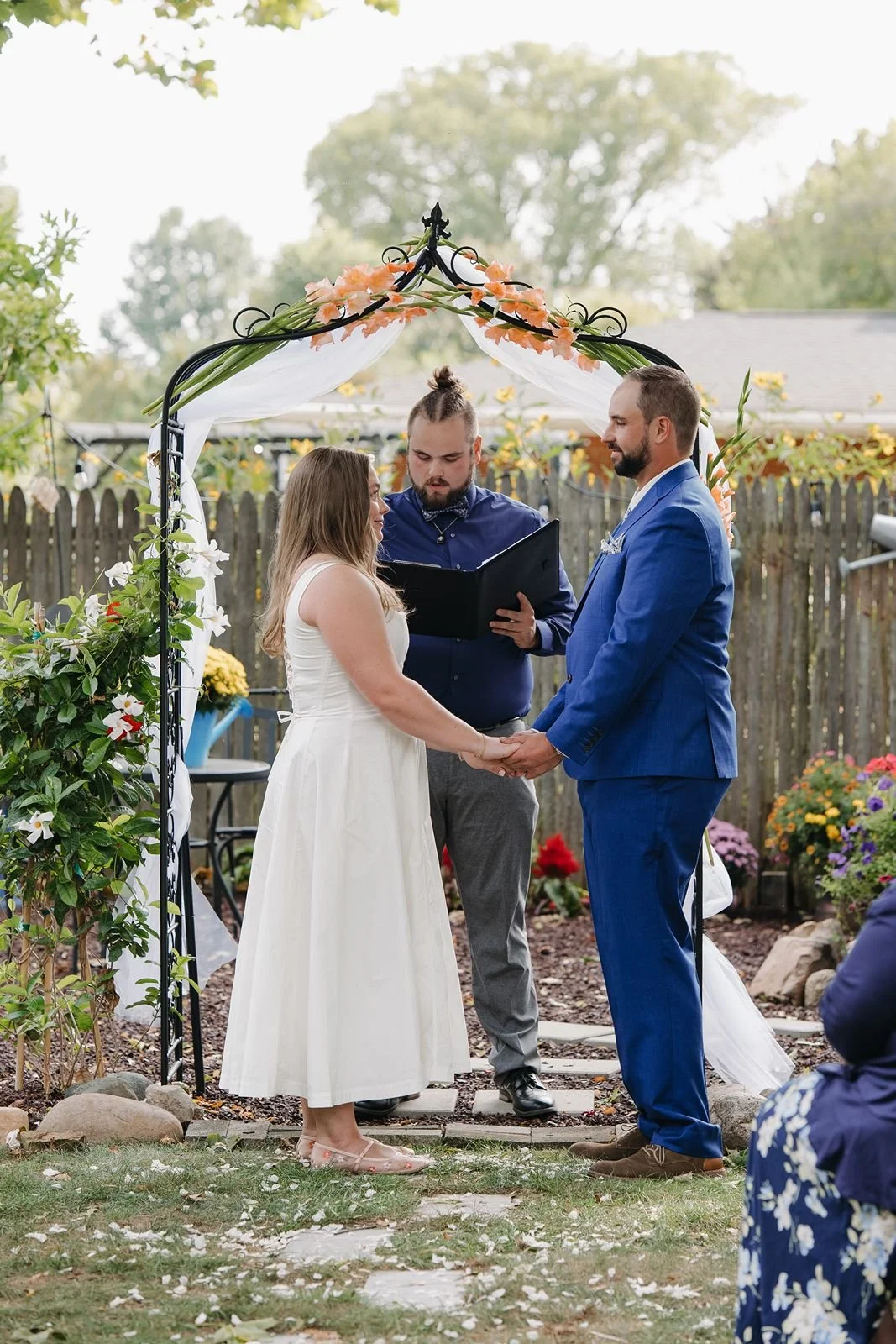 A bride and groom standing underneath an iron arch with florals woven in. They are standing in a backyard getting married.