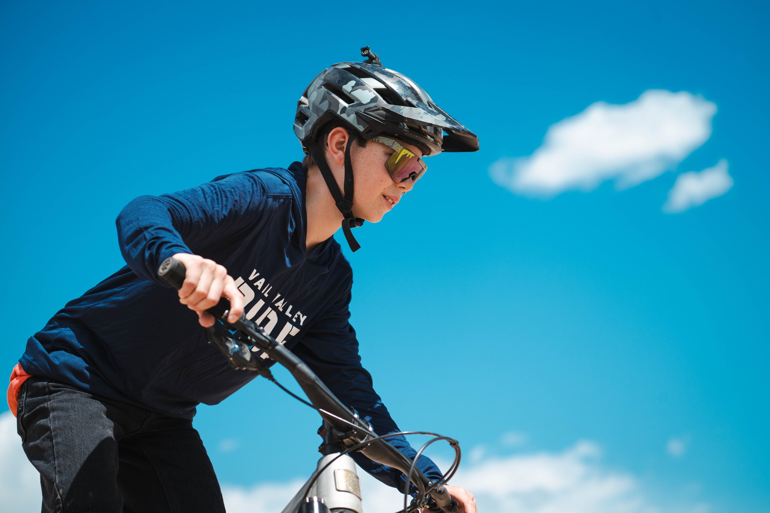 Group of mountain bikers, including children and adults, gathered outdoors with bikes, wearing helmets and gear, in a sunny area near houses and mountainous terrain.