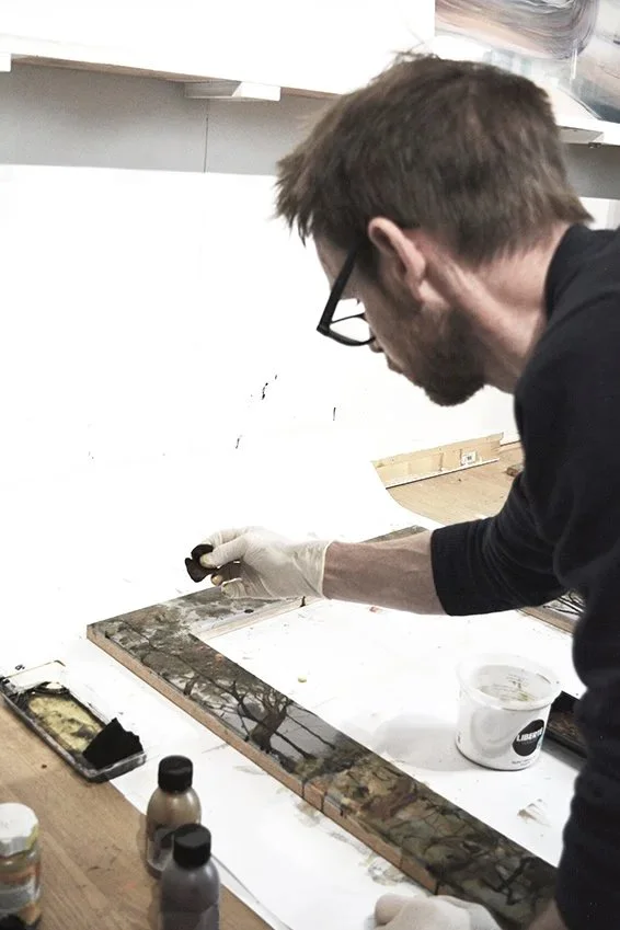 A person wearing glasses and gloves working on a painting or restoration project at a worktable, with paints and tools nearby.