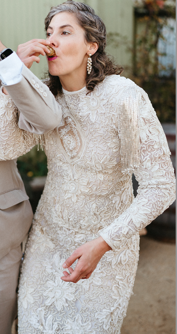A woman in an ornate wedding dress with lace details is being fed a piece of food, possibly cake, by another person. She is outdoors, wearing pearl earrings, and appears to be at a celebration or wedding.