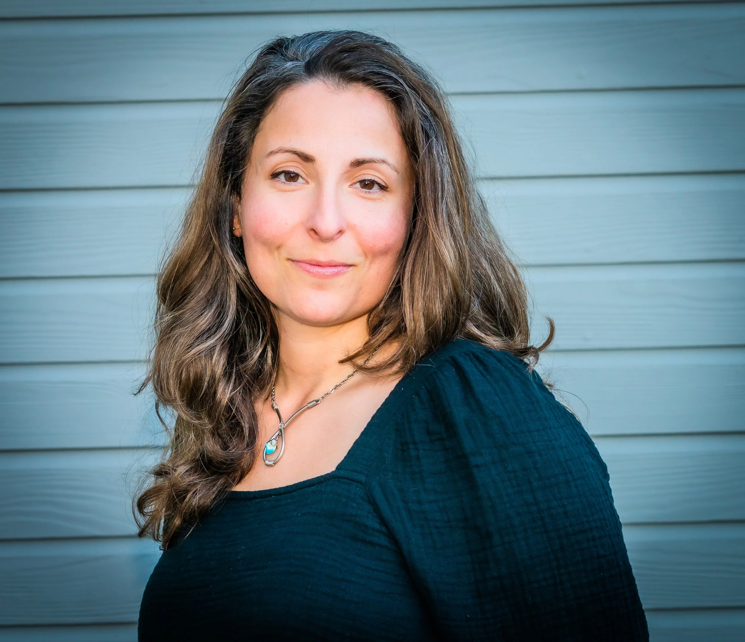 A woman with long wavy brown hair wearing a black top and silver necklace, standing in front of a gray wooden wall.
