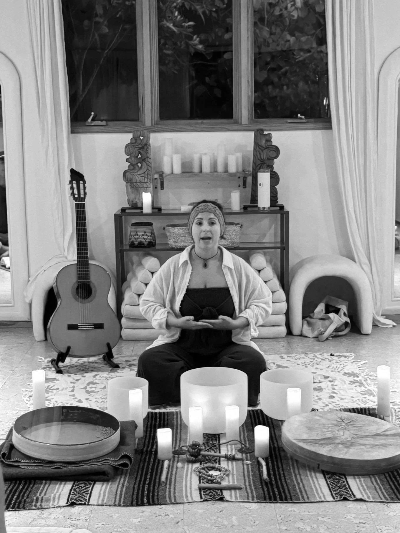 Woman sitting cross-legged on a cloth with candles, bowls, and spiritual objects, in front of a decorated wall with a guitar nearby.