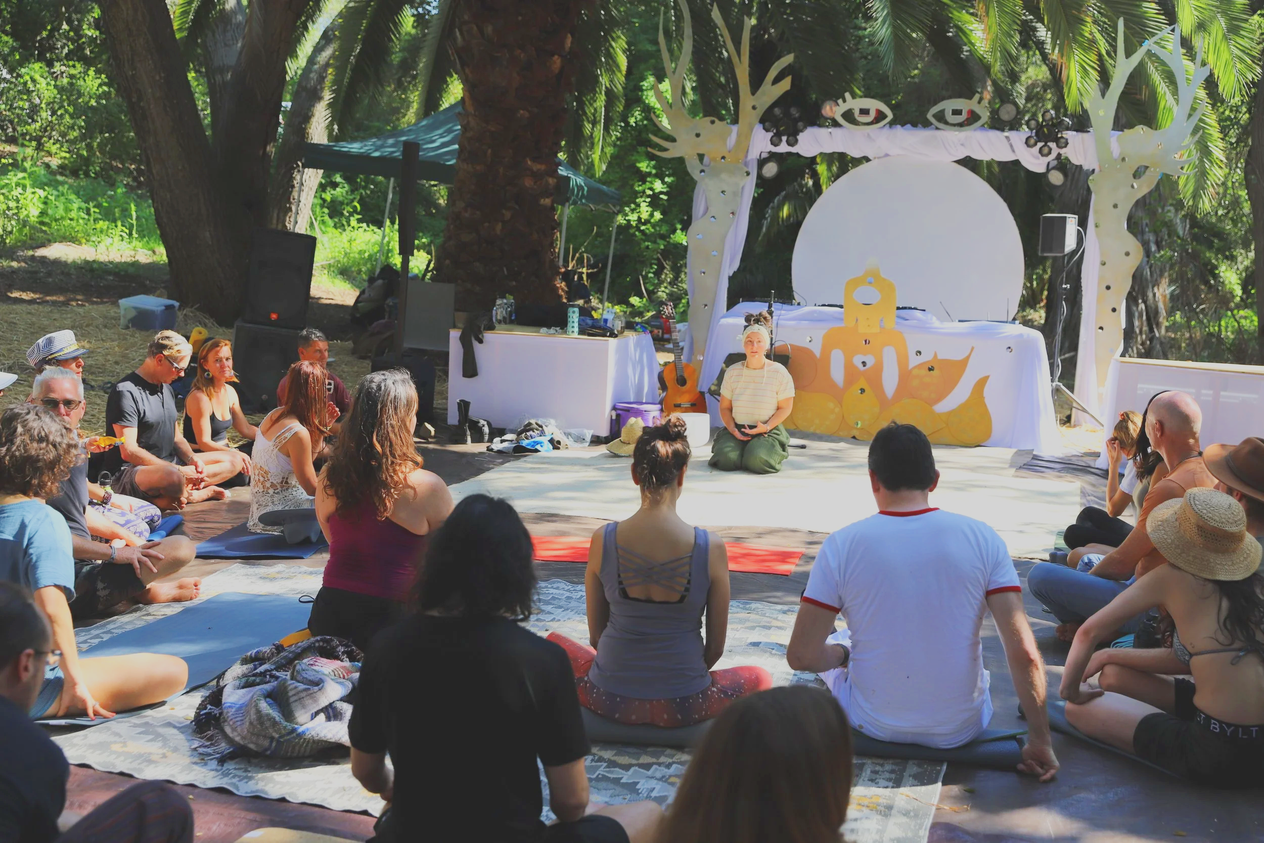 A group of people seated on mats and blankets outdoors, attending a meditation or yoga session led by a woman in front of a decorated stage with artistic designs and trees in the background.