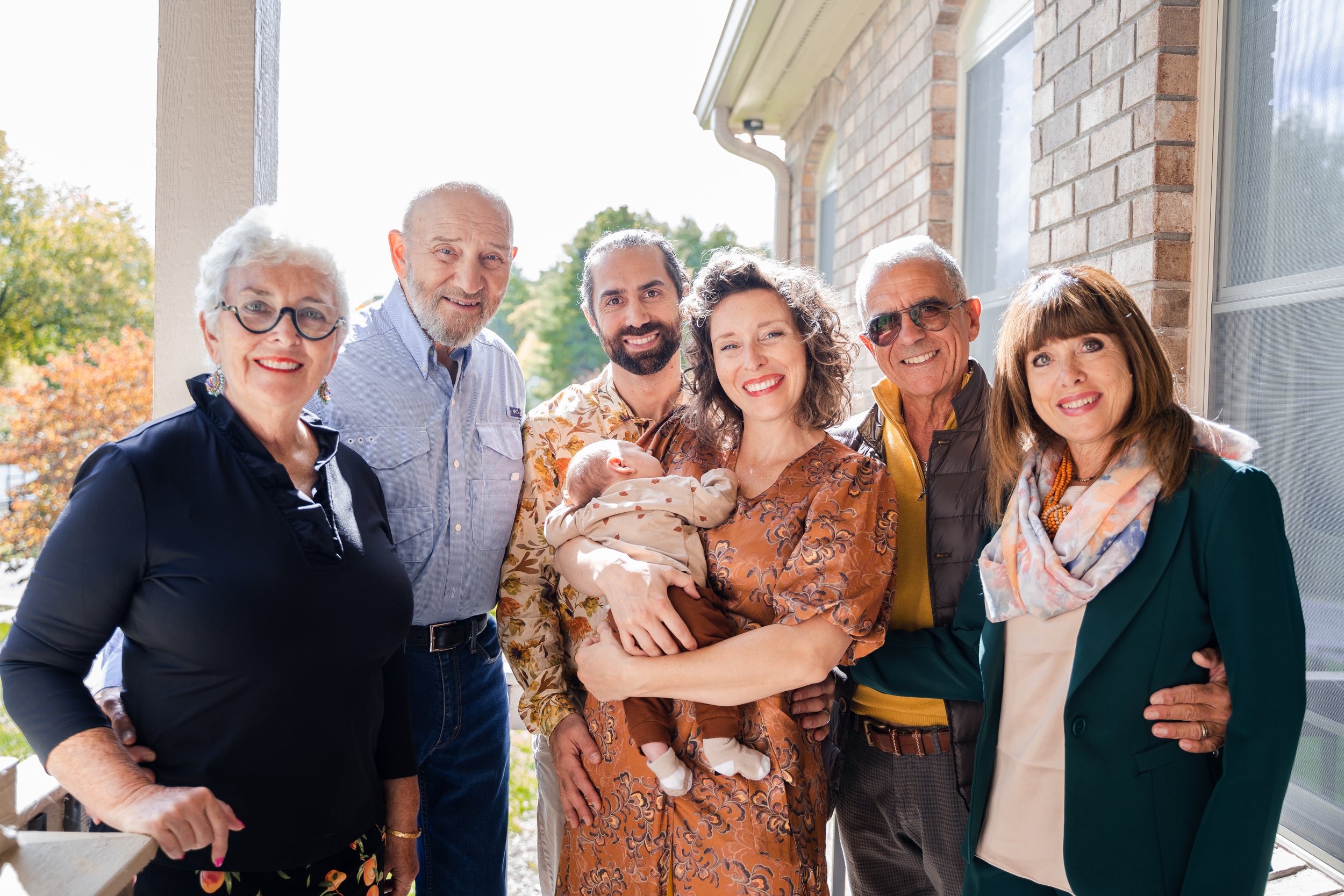 Family gathering outside on a porch, with grandparents, parents, and a baby, all smiling and dressed in casual and semi-formal clothing on a sunny day.
