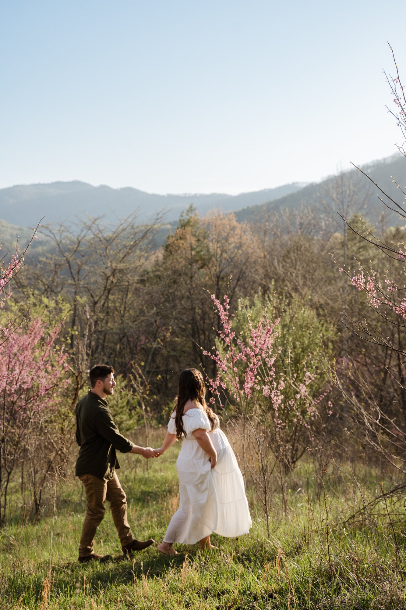 A man and pregnant woman hold hands while standing in a field with pink flowering trees and mountains in the background.