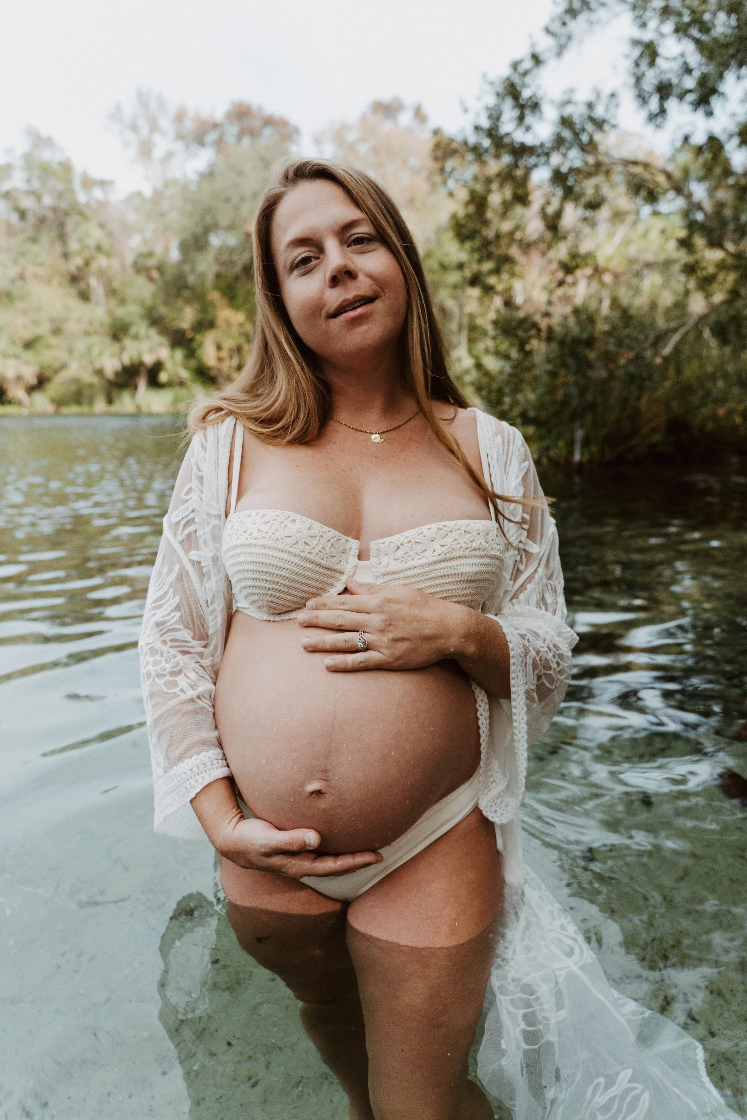 Pregnant woman standing in a river, holding her belly with one hand, wearing a white crochet top and a lace cover-up with a peaceful expression, surrounded by trees and nature.